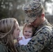 A U.S. Marine in full digital camouflage uniform and cover (cap) embraces a young toddler while a woman, likely his spouse or partner, leans in closely. The toddler is smiling broadly, suggesting a joyful reunion scene, possibly a homecoming from deployment. The emotional moment is captured outdoors with a blurred natural background.