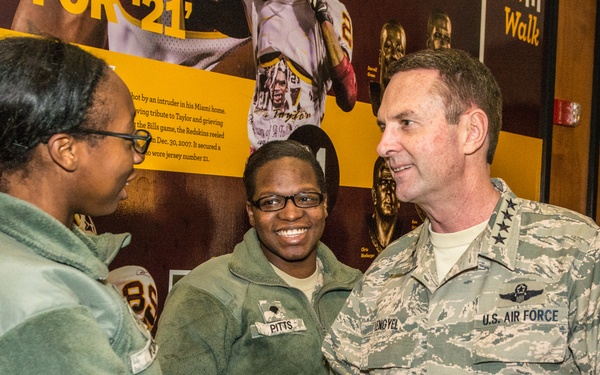 A U.S. Air Force officer in ABU (Airman Battle Uniform) is shaking hands with a young woman in civilian clothing, while another uniformed service member named 'Pitts' stands smiling in the background. The setting appears to be inside a Washington Redskins (NFL) museum or exhibit space, with team memorabilia and informational displays visible on the walls. The interaction appears to be a formal greeting or meet-and-greet event.