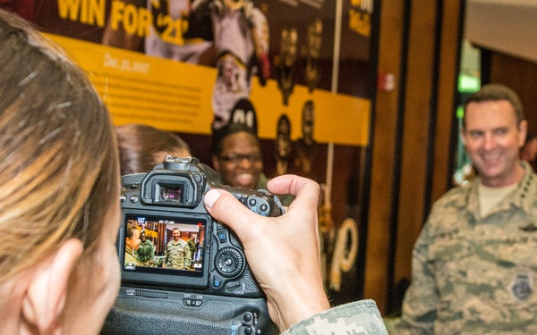 A female soldier in Army Combat Uniform (ACU) is photographing two military personnel — a smiling male in Air Force ABU (Airman Battle Uniform) and a civilian woman — at what appears to be an indoor military or sports-related event. The photographer is using a Canon DSLR camera mounted on a tripod head, and the subjects are visible both in the background and reflected on the camera's LCD screen. An American flag patch is visible on the photographer's sleeve.