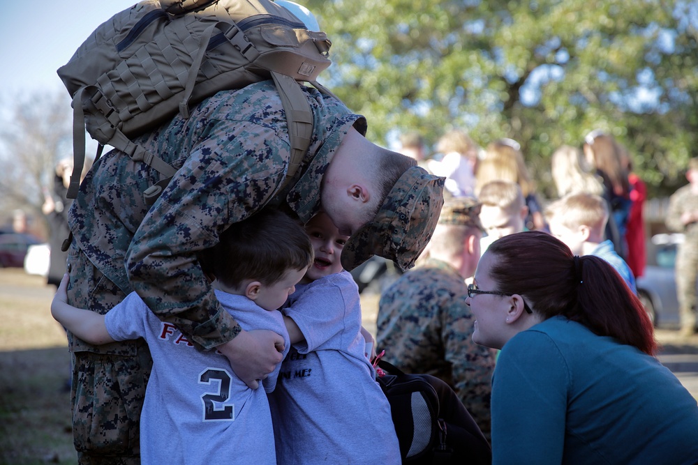 A U.S. Marine in digital camouflage (MARPAT) uniform with a large tactical backpack bends down to embrace two young children in an emotional reunion scene. A woman with dark red hair and glasses, likely the spouse or partner, kneels nearby watching the moment. The background shows additional Marines and civilians in a similar outdoor reunion setting.