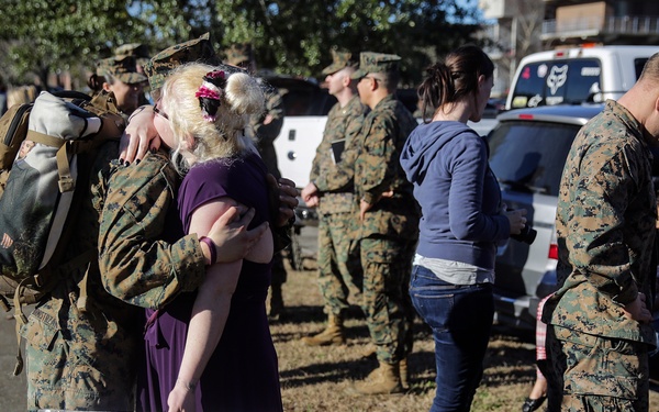 A military homecoming scene showing U.S. Marines in camouflage uniforms reuniting with their families. In the foreground, a woman with blonde hair in a purple dress embraces a Marine while holding a handmade countdown sign. The sign details the exact duration of the separation in years, months, weeks, days, hours, minutes, and seconds, ending with 'ONE KISS I'll forget them all!!'