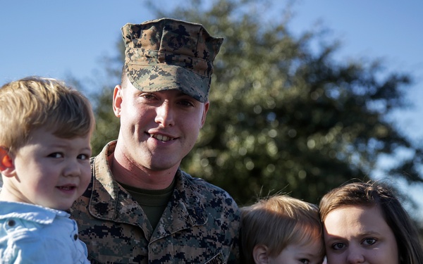 A U.S. Marine in digital camouflage uniform poses with what appears to be his family, including two young children and a woman, in an outdoor setting. The Marine is holding one child while the other child and the woman stand close beside him. The group appears to be smiling and in good spirits, suggesting a reunion or family gathering.