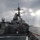 170124-N-JI086-068 The image shows a U.S. Navy guided-missile destroyer underway at sea, photographed from the deck of another vessel. The ship displays characteristic Arleigh Burke-class DDG features including a phased array radar mast, Harpoon missile launchers, and standard gray naval paint scheme. The scene is set under dramatic overcast skies with rays of sunlight breaking through the clouds.