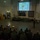 HMH-464 Marines attend Road to War Brief during Exercise Frigid Condor A military briefing is taking place in a large indoor facility where a uniformed presenter stands before a projected slide titled 'Regional Overview' showing a map labeled 'Canuckistan' overlaid on Canada. A large group of military personnel in camouflage uniforms are seated facing the presenter and projection screen. The setting appears to be a military training or operations facility, identifiable by unit insignia on the wall and restricted area signage.