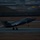 Lightning II strikes Iwakuni, F-35B arrives A fifth-generation stealth fighter jet, identifiable as an F-35, is captured during a nighttime takeoff or landing roll on a runway. The image was taken using a panning technique, resulting in a sharp subject against a motion-blurred background. Green formation lights and landing gear are clearly visible on the aircraft.