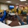 NPASE COMREL A person in U.S. Navy uniform is presenting to a group of elementary-aged children in a colorful classroom setting. Several children have their hands raised enthusiastically in response to the presenter. A television screen behind the presenter displays what appears to be a military helicopter over a naval vessel.