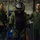 From bolts to bombs Three individuals are visible in what appears to be a military or naval vessel interior. The central figure is wearing a full EOD (Explosive Ordnance Disposal) bomb disposal suit with a large helmet and visor, flanked by two personnel in olive green flight suits. The setting appears to be a training or demonstration scenario aboard a ship.
