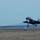 Lightning II strikes Iwakuni, F-35B arrives A fifth-generation stealth fighter jet, consistent with an F-35 Lightning II, is captured during a landing or takeoff roll on a military airstrip. The aircraft's landing gear is fully deployed and its nose gear landing light is illuminated. The setting appears to be dusk or dawn, with mountains visible in the background and runway edge lights visible along the tarmac.