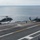 170118-N-NV908-021 Two U.S. Navy helicopters, identifiable as MH-60 Seahawks (numbered 20 and 2), are operating on the flight deck of an aircraft carrier at sea. One helicopter is in the process of lifting off or landing while the other remains on deck with rotors spinning. Flight deck crew members in colored jerseys are visible directing operations.