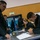 170119-N-OY799-004 A group of individuals in matching black fleece jackets and some in Navy digital camouflage uniforms are gathered around a metal table in what appears to be a military facility. Two people at the table are actively writing on documents using yellow highlighters or markers, while others stand and observe. The setting suggests an administrative check-in or sign-in process.