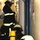 161229-N-N0901-780 The image shows multiple firefighters in full protective gear, including helmets and high-visibility black-and-yellow uniforms, operating in a narrow corridor or passageway. At least two firefighters are equipped with Dräger brand self-contained breathing apparatus (SCBA) with yellow compressed air cylinders on their backs. The setting appears to be an industrial or maritime interior, possibly during a training exercise or active emergency response.