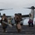 Marines return to USS Mesa Verde Military personnel, appearing to be U.S. Marines, are disembarking or moving across the flight deck of a naval vessel near a V-22 Osprey tiltrotor aircraft. The Marines are carrying gear, backpacks, and equipment including what appears to be a riot shield and a fuel/water jerry can. Flight deck crew in colored vests and helmets are visible managing operations in the background.