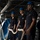 The aircraft carrier is deploying as part of the George H.W. Bush Carrier Strike Group in support of maritime security operations and theater security cooperation efforts in the U.S. 5th and 6th Fleet areas of operations Several naval personnel in dark blue uniforms and blue hard hats are handling large mooring lines aboard what appears to be a military vessel. Three sailors in the foreground are holding a thick braided rope, likely during a mooring or line-handling evolution. The setting is an interior deck area of a ship with visible overhead piping and industrial infrastructure.