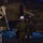 Lightning II strikes Iwakuni, F-35B arrives A nighttime scene on a flight line showing two personnel in flight suits standing near a military jet aircraft. The aircraft is marked with 'VMFA-121,' identifying it as belonging to Marine Fighter Attack Squadron 121. One individual is wearing a full flight helmet with visor equipment, suggesting a pilot conducting pre-flight or post-flight procedures.