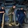 170118-N-SK327-127 Military personnel are working together aboard what appears to be a naval vessel, handling large mooring lines or ropes in an interior deck space. The group includes at least one Marine in woodland MARPAT camouflage and two Navy sailors in dark blue coveralls and Navy ball caps. They appear to be coiling, moving, or stowing heavy rope lines in a below-deck area surrounded by pipes and cables.