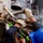 USS Lake Champlain (CG 57) Replenishment-at-Sea Workers in protective gear are handling heavy rigging hardware, likely shackles or chain links, on what appears to be a naval vessel or large industrial ship. At least two workers are visible, one wearing green tactical gloves and another wearing tan leather work gloves, along with a blue hard hat. The equipment shows significant rust and wear, indicating aged maritime hardware.