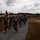 MWCS-28 Spartan Cup A large formation of U.S. Marines in olive drab PT (physical training) shirts and camouflage trousers conducts a unit run along a paved path. Two guidon bearers carry red Marine Corps flags at the front of the formation. The setting appears to be a military installation, likely on the U.S. East Coast based on the flat terrain and pine tree vegetation.