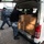 USS Bonhomme Richard (LHD 6) Sailors participate in a Food Drive Two individuals wearing U.S. Navy Working Uniform (NWU) digital camouflage are loading or unloading cardboard boxes from the rear of a white van in a parking lot. One person is actively handling the stacked boxes while the other approaches from behind. The setting appears to be a military installation or base facility based on the uniforms and surrounding buildings.
