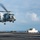 170123-N-BL637-018 A military helicopter, appearing to be an MH-60 Seahawk, is hovering low over the flight deck of a naval aircraft carrier. A deck crew member wearing a green jersey and brown float coat is signaling with outstretched arms, guiding the helicopter's landing. The scene takes place at sea under partly cloudy skies.