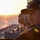 MKI Captain A military naval officer stands on the deck of a warship during sunrise or sunset, gazing toward the horizon over open water. He is wearing a dark Navy ball cap with gold insignia and an American flag patch, along with a dark jacket bearing military patches. The dramatic backlighting creates a silhouette-like effect with warm golden tones.