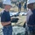 170120-N-AW702-002 Two U.S. Navy personnel in the foreground, wearing blue digital camouflage uniforms, white hard hats, and black gloves, appear to be in conversation during what looks like a field exercise or equipment setup operation. Large tarpaulins or tent materials are spread across the ground around them. Numerous other military personnel in similar uniforms are visible in the background engaged in the same activity.