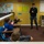 NPASE COMREL A person in U.S. Navy camouflage uniform is presenting or speaking to a small group of young children seated at school desks in a colorful elementary classroom. One child has their hand raised, suggesting active participation or a question-and-answer session. The classroom features educational posters including a world map, cursive/manuscript alphabet chart, and various learning materials.