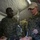 2d Medical Bn Field Exercise Two U.S. Navy personnel in Marine Corps digital camouflage uniforms are conducting what appears to be a field medical training exercise inside a military tent. One personnel holds an IV bag over a simulated casualty lying on a stretcher, while the other, wearing an 'INSTRUCTOR' cap and holding a clipboard, observes and evaluates. The patient has simulated wounds including blood makeup on the face and is fitted with a breathing tube/airway device.