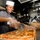 USS Lake Champlain (CG 57) Galley A smiling chef in a white toque and gray uniform is cutting a large sheet pan of pepperoni pizza using a pizza wheel cutter in a commercial kitchen. She is wearing plastic gloves and appears to be in a military or institutional galley setting. A second person in similar attire is visible working in the background.