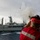 USS Lake Champlain (CG 57) Replenishment-at-Sea A naval personnel member wearing a red helmet labeled 'GUNNER' and a red safety vest is firing a line-throwing gun toward a large gray naval replenishment vessel. A projectile and red line are visible in mid-air, trailing toward the ship. Smoke is visible rising from the area near the ship, consistent with a line-throwing operation during an underway replenishment (UNREP) evolution at sea.