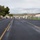 Camp Pendleton flood 2017 A wide multi-lane road is partially blocked by orange traffic cones and white barricades in the middle distance, suggesting a checkpoint or road closure. The road appears wet, indicating recent rainfall, and is flanked by trees on the left and industrial or institutional buildings on the right. The setting appears to be a semi-rural or base/facility environment with rolling green hills in the background.