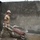 NMCB 5 Classroom Construction Project in Panay A construction worker wearing military-style camouflage pants, a tan t-shirt, and a white hard hat is pushing a red wheelbarrow containing wet concrete or mortar. He is working inside an unfinished concrete block building with freshly plastered walls. The setting appears to be a construction or humanitarian aid project.