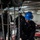 170118-N-TV230-188 A military sailor wearing a blue hard hat and safety goggles uses a mallet or hammer to work on heavy anchor chain equipment in what appears to be the chain locker or anchor handling area of a naval vessel. Two other personnel in dark coveralls and hard hats are visible in the background observing or assisting. The environment is an enclosed, industrial shipboard space with overhead chain hoists and heavy mechanical equipment.