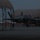 Lightning II strikes Iwakuni, F-35B arrives A military stealth fighter jet, consistent with an F-35 variant, is positioned on a flight line apron between dome-shaped aircraft shelters (hardened aircraft shelters or portable canopy structures) during dusk or dawn conditions. Ground crew personnel are visible near the aircraft. Navigation lights (green and red) are illuminated on the aircraft and surrounding area.
