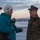 Boots in the Snow A journalist or reporter is conducting an outdoor interview with a military personnel wearing MARPAT (Marine Corps digital camouflage) uniform and cover. The interview is taking place in a cold, wintry environment, likely at an airfield or open tarmac area during dawn or dusk. A professional video camera on a tripod is visible in the background left.