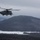 HMH-464 Marines Conduct Flight Operations during Exercise Frigid Condor A large military helicopter, consistent with a CH-53E Super Stallion, is flying over a snow-covered mountainous landscape under overcast winter skies. The helicopter appears to be in active flight with rotor blades visibly spinning. The terrain below features forested hills, open snow-covered fields, and rolling mountains in the background.