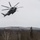 HMH-464 Marines Conduct Flight Operations during Exercise Frigid Condor A large military helicopter, consistent with a Sikorsky CH-53E Super Stallion, flies low over a snow-covered clearing in a forested, hilly winter landscape. The rotor blades are motion-blurred, indicating active flight. The terrain features mixed deciduous and coniferous trees with snow-dusted hills in the background under an overcast sky.