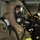 161229-N-N0901-775 A person wearing full firefighting or emergency response protective gear, including a full-face respirator/gas mask and a tan/brown protective suit with a 'Body-Guard 2000' patch, stands in what appears to be a ship's interior corridor. Several other similarly equipped personnel are visible in the background. The setting and equipment suggest a damage control or firefighting drill, likely aboard a naval vessel.