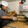 USS Lake Champlain (CG 57) Galley A female chef wearing a white toque and dark chef's coat is working in a commercial kitchen, handling what appears to be cooked noodles or spaghetti using gloved hands. She is transferring pasta from a large tray into a smaller metal pan. A prominent 'CLEAN AS YOU GO' sign is visible on stainless steel equipment to her right.