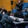 170119-N-OY799-015 A military briefing or meeting is taking place in what appears to be a gymnasium or auditorium with blue bleacher seating. A senior military personnel member is standing and speaking into a microphone while addressing a group of seated service members wearing Navy Working Uniform (NWU) camouflage pattern. The attendees are seated at tables and in bleachers, attentively listening to the speaker.