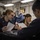 170123-N-MJ135-061 Several military personnel in dark navy uniforms are gathered around a table in what appears to be a shipboard mess or briefing area. Two individuals in the foreground are reviewing printed documents, while others stand or sit nearby. The setting features industrial overhead lighting and exposed piping consistent with a naval vessel interior.