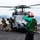 170117-N-HD638-009 A military helicopter, appearing to be an MH-60 Seahawk, is on the flight deck of a naval vessel with rotor blades spinning. A deck crew member in a green shirt and red helmet is giving hand signals to the cockpit, while armed personnel in tactical gear stand nearby. The scene takes place on open water with mountainous islands visible in the background.