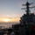 170117-N-PP996-015 A U.S. Navy guided-missile destroyer is underway at sea during sunset or sunrise, with crew members visible on the deck in safety gear. The ship's superstructure, mast, radar systems, and forward gun mount are prominently visible in silhouette against the warm horizon light. Multiple sailors wearing hard hats and life vests are gathered on the forward deck, suggesting an active evolution or drill.