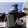 USS Lake Champlain (CG 57) Crew-Served Weapons Shoot Two military personnel are positioned on the deck of a naval vessel at sea. The foreground figure, wearing a helmet, face covering, gloves, and body armor marked 'NAVY', is manning a M60 or M240 machine gun with an M19A1 ammunition can attached. A second person in the background wears a dark uniform and communications headset while observing the water.