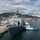 USS Bonhomme Richard (LHD 6) USS Ashland Departs A large gray U.S. Navy warship bearing hull number 46 is docked at a pier, assisted by a blue tugboat marked with the letter 'S'. The scene appears to be at a naval base in Japan, identifiable by the surrounding cityscape, mountainous terrain, and harbor infrastructure. A second white vessel is visible in the background on the water.