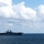 COMPTUEX A large amphibious assault ship is underway on open ocean under a partly cloudy sky. Multiple helicopters are visible, with at least one airborne to the port side of the vessel and several aircraft spotted on the flight deck. The ship appears to be conducting flight operations in calm sea conditions.