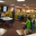 NPASE COMREL Two individuals in military uniforms (appearing to be U.S. Navy camouflage pattern) are presenting to a group of children and adults in a colorful classroom setting. One uniformed person is speaking near a wall-mounted TV displaying images, while children sit at desks attentively. The setting appears to be an elementary school or educational facility with bright yellow walls and child-sized furniture.