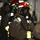 161229-N-N0901-777 The image shows a group of firefighters or emergency responders in full protective gear, including SCBA (Self-Contained Breathing Apparatus) masks, helmets with mounted lights, and tan/brown turnout gear with yellow reflective strips. They appear to be inside a confined industrial or naval vessel space during what looks like a training exercise or active emergency response. The lead figure is wearing a full-face respirator mask and is looking directly at the camera.