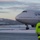 Boots in the Snow A Military Police officer stands on a snow-covered airport tarmac observing a United Airlines Boeing 747 aircraft taxiing or parked nearby. The officer is wearing a high-visibility yellow vest with 'MILITARY POLICE' printed on the back and a red beret, and is holding a radio. The scene appears to be at dusk or dawn in a cold, wintry environment with snow-covered ground and mountains visible in the background.
