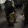 From bolts to bombs A person wearing a full EOD (Explosive Ordnance Disposal) bomb suit with helmet and face shield is carrying a green military-style container or case in what appears to be the interior of a naval vessel or military facility. A second individual in camouflage uniform stands nearby observing. The scene appears to be either a training exercise or an operational procedure.