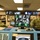 170123-N-TI693-111 Three military personnel in Navy Working Uniform (NWU) camouflage are seated at a control console monitoring multiple CCTV feeds. The central Dell monitor displays a 9-panel grid of security camera footage showing what appears to be industrial or mechanical spaces. The facility appears to be a military monitoring or control room with multiple display screens along the wall.